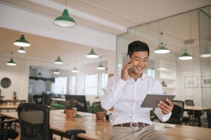 Smiling young Asian businessman on the mobile phone sitting on a desk